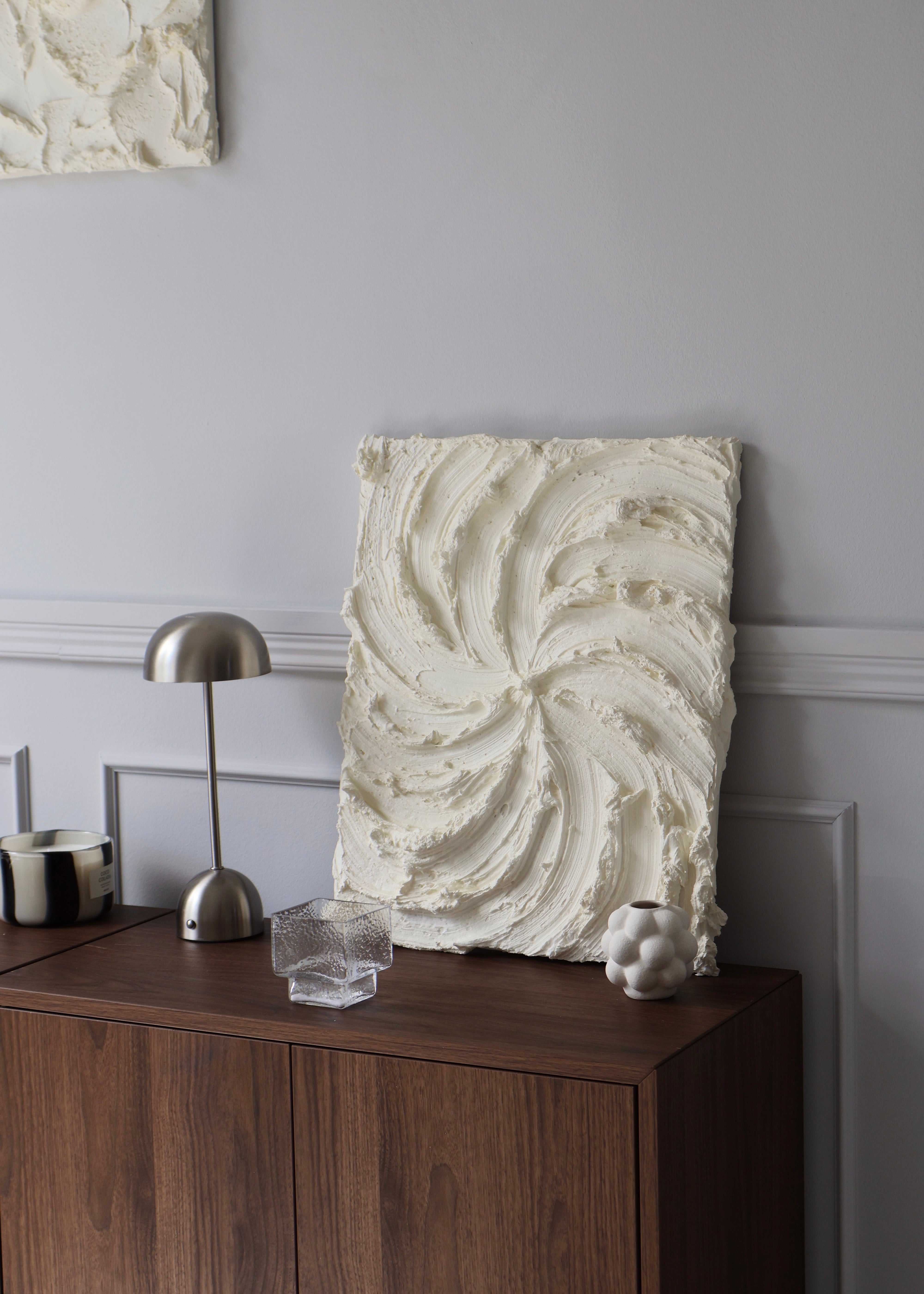 An off-white textured spiral wall art piece leaning against a gray wall on a dark wood cabinet, styled with a silver table lamp, a glass, and a small decorative object.